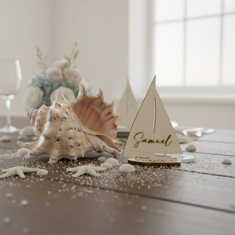 Puede incluir: Un tarjetero de madera con forma de velero con el nombre "Samuel" grabado. Una gran concha y estrellas de mar est&aacute;n sobre una mesa de madera, con arena esparcida y peque&ntilde;as piedras blancas. Una copa de vino y flores est&aacute;n en el fondo.