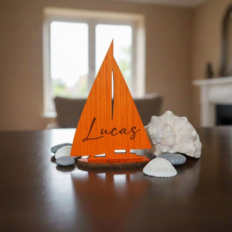 Puede incluir: Un velero de madera naranja con el nombre "Lucas" grabado en la vela. El velero est&aacute; sobre una base de madera y est&aacute; rodeado de peque&ntilde;as piedras, una concha y una caracola. Es un art&iacute;culo decorativo.