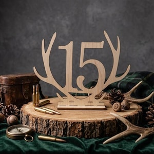 May include: A wooden table centerpiece with the number 15, framed by antlers. The centerpiece sits on a wood slice, surrounded by pine cones, bullets, and a compass. The background includes a green plaid blanket and a dark, textured wall.