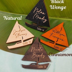 Puede incluir: Tarjetas de mesa de madera con forma de velero en acabados natural, negro, naranja y chocolate. Cada tarjeta presenta un nombre y un número de mesa. La imagen incluye el texto "Natural", "Black Wenge", "Cinnamon" y "Chocolate".