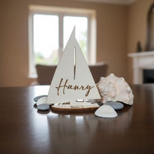 Puede incluir: Un tarjetero de madera con forma de velero, con el nombre "Harry" grabado en la vela. La base indica "Table 3". Conchas marinas y guijarros complementan la escena, creando un tema n&aacute;utico. El velero es de madera clara.