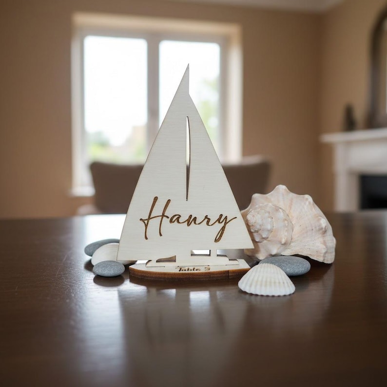 Puede incluir: Un tarjetero de madera con forma de velero, con el nombre "Harry" grabado en la vela. La base indica "Table 3". Conchas marinas y guijarros complementan la escena, creando un tema n&aacute;utico. El velero es de madera clara.