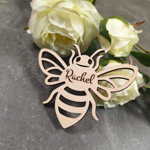 May include: A wooden bee-shaped cutout with the name "Rachel" engraved on it. The bee has detailed wings and antennae. The background features white flowers and a gray surface.