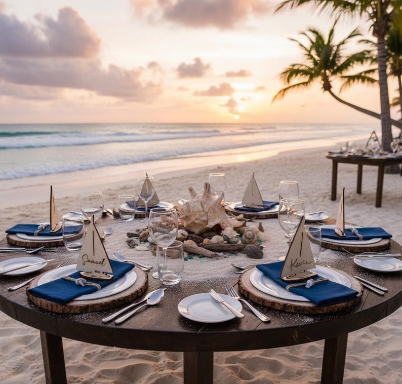 Puede incluir: Una mesa redonda de madera preparada para un evento en la playa. Cada cubierto incluye un plato blanco, una servilleta azul, cubiertos y una tarjeta de lugar con forma de velero de madera. La mesa est&aacute; decorada con conchas marinas y madera flotante, con una puesta de sol al fondo.