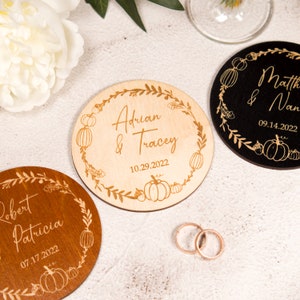 Könnte beinhalten: Drei runde Holz-Untersetzer für die Hochzeit mit Herbst-Motiv. Die Untersetzer zeigen einen Kranz aus Blättern und Kürbissen mit den Namen des Paares und dem Hochzeitsdatum. Die Untersetzer sind in Naturholz, Schwarz und Braun.