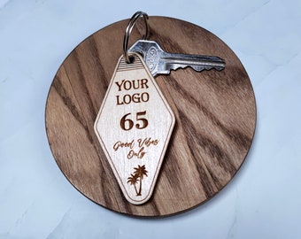 Op de afbeelding: Een houten sleutelhanger in de vorm van een diamant, met de tekst "YOUR LOGO", "65" en "Good Vibes Only" met een palmboomgrafiek. De sleutelhanger is bevestigd aan een zilveren sleutelring met een zilveren sleutel.