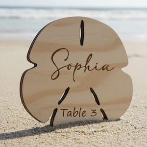 May include: A wooden sand dollar-shaped table sign with the name "Sophia" in cursive and "Table 3" engraved. The sign is light brown and sits on a sandy beach with the ocean in the background.