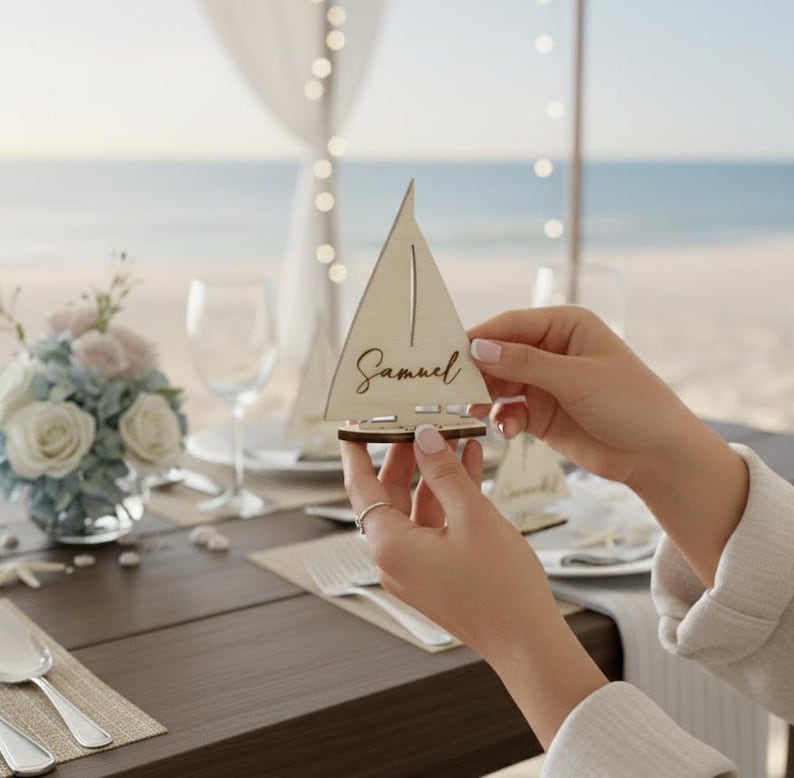 May include: A wooden sailboat place card with the name "Samuel" engraved on the sail. The place card is held by a person. The background includes a table setting with flowers and a beach view.
