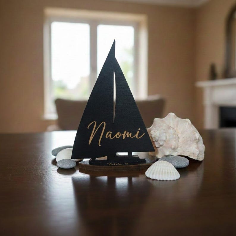 Puede incluir: Decoraci&oacute;n de mesa en forma de velero negro con el nombre "Naomi" en escritura dorada. El velero se asienta sobre una base de madera, con peque&ntilde;as piedras y conchas marinas cerca. La mesa es marr&oacute;n oscuro.
