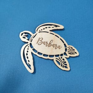 May include: A wooden cutout of a sea turtle with the name "Barbara" engraved on the shell. The turtle is a light brown color and has a detailed design.