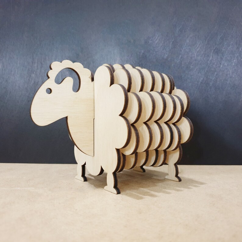 May include: Wooden sheep-shaped coaster set. The coaster is made of multiple layers of wood, creating a 3D effect. The sheep has a brown outline and a natural wood colour.