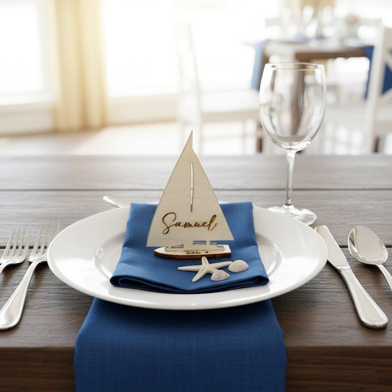 Puede incluir: Una tarjeta de lugar de madera con forma de velero con el nombre "Samuel" sobre una servilleta azul, colocada en un plato blanco. Una estrella de mar y una concha acent&uacute;an el tema n&aacute;utico. Cubiertos y copa de vino completan la mesa.