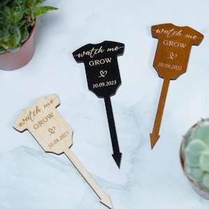 Könnte beinhalten: Drei hölzerne Pflanzenschilder in Form von Baby-Bodys in Natur-, Schwarz- und Brauntönen. Jeder Marker trägt die Worte "watch me GROW", ein Herzsymbol und das Datum "10.09.2023" eingraviert.