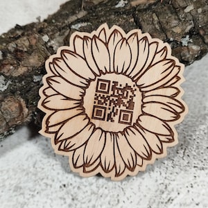 Könnte beinhalten: Ein hellfarbiger Holzausschnitt in Form einer Sonnenblume mit einem dunkelbraunen, lasergravierten QR-Code in der Mitte. Die Blütenblätter sind mit feinen, dunkelbraunen Linien detailliert und das Design ist flach mit einer glatten Holzmaserung.