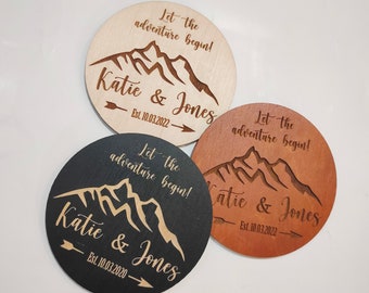 May include: Three round wooden coasters featuring a mountain range design and the text "Let the adventure begin!"  "Katie & Jones" and the date "Est. 10.03.2022". The coasters are in natural wood, black, and brown.