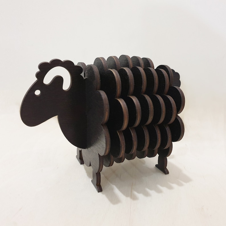 May include: A black wooden sheep-shaped coaster with a white ear. The coaster is made of multiple layers of wood, creating a 3D effect.