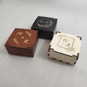 Pode incluir: Três caixas quadradas de madeira com desenhos gravados. Uma é marrom escuro com "M & R", outra é preta com "B + K" e a terceira é de madeira clara com "D / A". Cada caixa tem detalhes nos cantos.