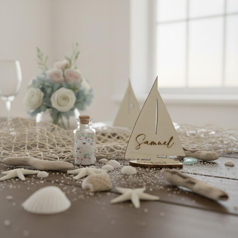 Puede incluir: Una decoraci&oacute;n de mesa de tem&aacute;tica n&aacute;utica con tarjetas de lugar de velero de madera, una botella de vidrio llena de confeti de colores, conchas marinas y madera flotante. Un ramo de flores blancas y azules se encuentra en el fondo. La tarjeta de lugar del velero tiene el nombre "Samuel".