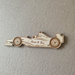 May include: Wooden save the date magnet shaped like a racing car with the text "Save the Date" and "Anna & Tom" with the date "12/04/2025" printed on it.