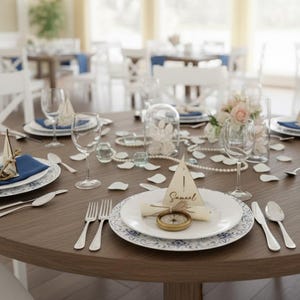 Puede incluir: Una mesa redonda de madera preparada para una boda o evento especial. La mesa est&aacute; adornada con platos blancos, servilletas azules, cubiertos, copas de vino y elementos decorativos como perlas, flores y una tarjeta de mesa con tem&aacute;tica de br&uacute;jula.