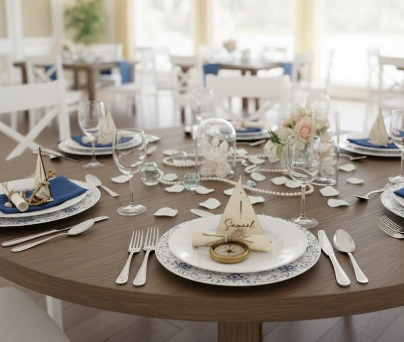 Puede incluir: Una mesa redonda de madera preparada para una boda o evento especial. La mesa est&aacute; adornada con platos blancos, servilletas azules, cubiertos, copas de vino y elementos decorativos como perlas, flores y una tarjeta de mesa con tem&aacute;tica de br&uacute;jula.