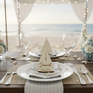 Puede incluir: Una mesa bellamente decorada con arreglos de mesa de tem&aacute;tica n&aacute;utica. Cada lugar incluye un plato blanco, una servilleta de lino, cubiertos y un portatarjetas de barco de vela de madera con el nombre "Samuel". La mesa est&aacute; decorada con conchas marinas y flores, con una playa de fondo.