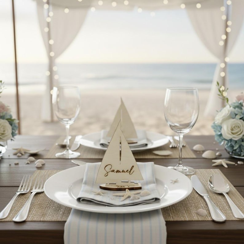 Puede incluir: Una mesa bellamente decorada con arreglos de mesa de tem&aacute;tica n&aacute;utica. Cada lugar incluye un plato blanco, una servilleta de lino, cubiertos y un portatarjetas de barco de vela de madera con el nombre "Samuel". La mesa est&aacute; decorada con conchas marinas y flores, con una playa de fondo.