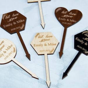 Puede incluir: Marcadores de jardín de madera en varias formas, incluyendo un hexágono, un círculo y un corazón. Cada marcador tiene el texto "let love grow" y los nombres "Emily & Mike" con una fecha. Los marcadores son en tonos marrón y negro.