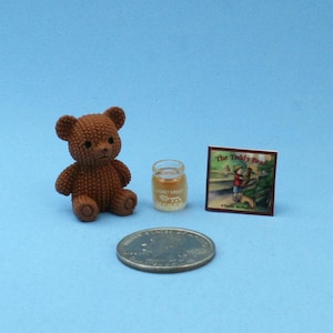 May include: A miniature brown textured teddy bear figurine, a small clear jar filled with a golden liquid, and a square book titled The Teddy Bear, all positioned next to a silver United States quarter coin for scale on a light blue surface.