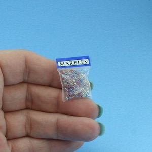 May include: A miniature clear plastic bag filled with tiny, multi-colored iridescent spheres. The top of the bag features a blue header strip with the word MARBLES printed in white capital letters. The bag is small enough to be held in a palm.