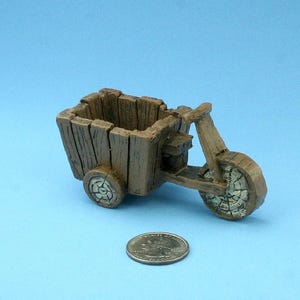 Puede incluir: Un triciclo de madera en miniatura con una jardinera rectangular. Las ruedas y la cesta tienen una textura de madera envejecida. Una moneda está al lado del triciclo para la escala. El fondo es azul claro liso.