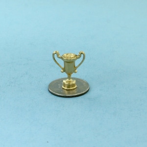 May include: A small, gold-colored trophy with two handles, set on a silver coin. The trophy is detailed with a cup-like design and a circular base. The background is a solid light blue.