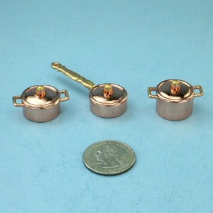 May include: Miniature copper pots with brass handles and lids. One pot has a long handle, and two have small side handles. A U.S. quarter is next to the pots for size comparison. The pots are on a light blue surface.