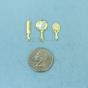 May include: A set of three gold-toned miniature hairdressing tools: a comb, a round hand mirror, and a hairbrush. The tools are arranged on a light blue surface next to a US quarter for scale.