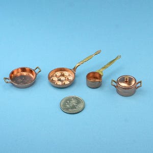 May include: Miniature copper cookware set including a frying pan, a saucepan, a ladle, and a pot with a lid. The set is displayed on a blue background with a quarter for size comparison. The cookware has brass handles.