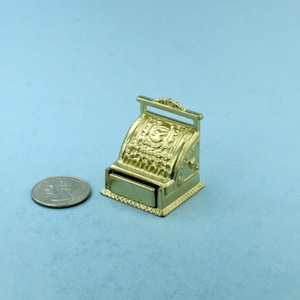 May include: A miniature gold-colored cash register with a drawer and a handle. The cash register is sitting on a light blue surface next to a silver coin.