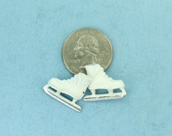 May include: Miniature white ice skates with silver blades are positioned near a quarter on a light blue background. The skates are detailed with laces and a classic design, suggesting a theme of winter sports or crafts.