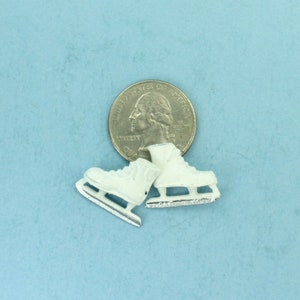 Puede incluir: Dos peque&ntilde;os patines de hielo blancos con cuchillas plateadas se encuentran junto a una moneda sobre un fondo azul claro. Los patines, con su dise&ntilde;o detallado, evocan deportes de invierno o proyectos de manualidades.