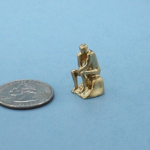 May include: A gold-colored figurine of a person sitting on a rock, similar in style to the famous "The Thinker" statue.