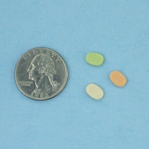 May include: Three oval-shaped candies in different colors: green, orange, and white. The candies are on a light blue surface next to a silver US quarter.