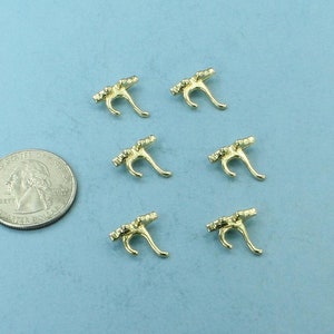 May include: Six small, gold-colored metal charms in a decorative shape, arranged on a light blue surface. A US quarter coin is next to the charms for size comparison. The charms have a curved, stylized design.