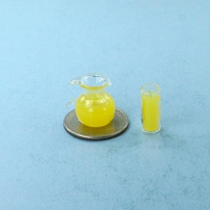 Puede incluir: Jarra de vidrio en miniatura y un vaso de líquido amarillo, posiblemente limonada, sobre una moneda. La jarra tiene asa y boquilla. El vaso es cilíndrico. Los artículos están sobre una superficie azul claro.