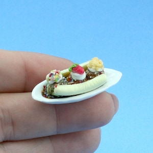 May include: A miniature banana split dessert served on a small white oval plate. The dish features three scoops of ice cream topped with whipped cream, a red strawberry, and colorful sprinkles, all resting on a sliced yellow banana base.