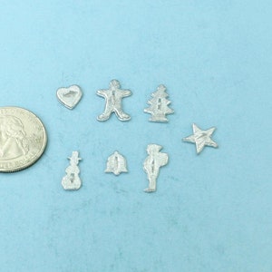 May include: A collection of silver-colored metal charms in the shape of a heart, a snowman, a Christmas tree, a star, a violin, and a bell. The charms are arranged on a light blue surface.