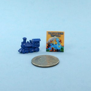 May include: A miniature blue toy train next to a small book with the title "The Little Engine That Could" and a quarter coin, all on a light blue surface. The train is a detailed model.