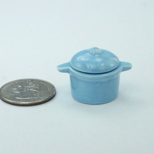 May include: A small, light blue ceramic pot with a lid, featuring two small handles. The pot is next to a quarter coin for size comparison. The pot is approximately 1 inch in diameter.