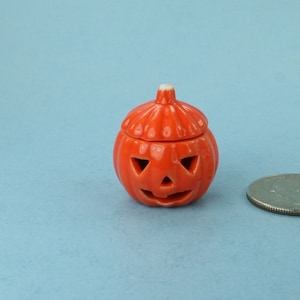 May include: A small orange ceramic pumpkin with a lid, featuring a carved jack-o'-lantern face. The pumpkin is about the size of a US quarter.