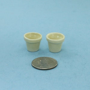 May include: Two small, off-white ceramic flower pots, approximately 2 inches in diameter, on a light blue background.