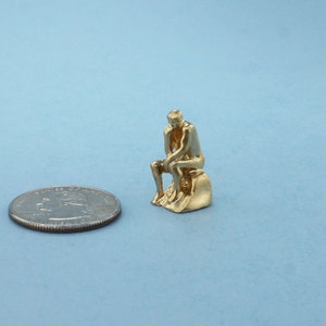May include: A gold-colored figurine of a person sitting and thinking, similar to the famous "The Thinker" statue. The figurine is about the size of a US quarter.