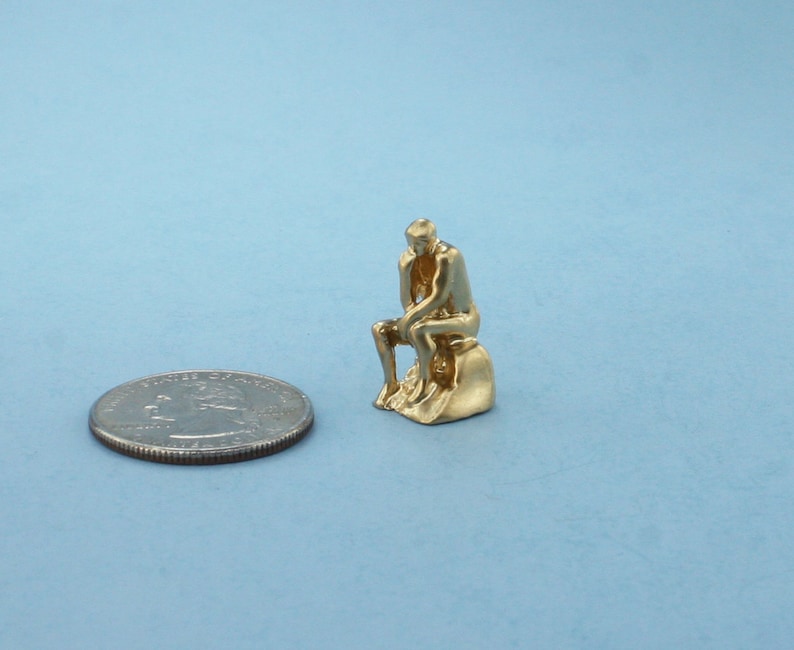 May include: A gold-colored figurine of a person sitting and thinking, similar to the famous "The Thinker" statue. The figurine is about the size of a US quarter.